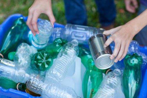 A close-up of a person’s hands sorting plastic waste into a blue recycling bin filled with various plastic bottles and containers. The bottles are made of clear and green plastic, with some having ridged or smooth surfaces, and are capped with different coloured lids. The person's left hand is holding a clear plastic bottle with a ridged neck, while the right hand is placing a silver tin can into the bin. The background shows a grassy area, indicating an outdoor setting, and the person is wearing blue jeans. The scene highlights waste separation and recycling efforts commonly associated with rubbish removal services, such as those offered by Tottenham House Clearance, within the Tottenham area, and is set against the context of environmentally conscious waste disposal.