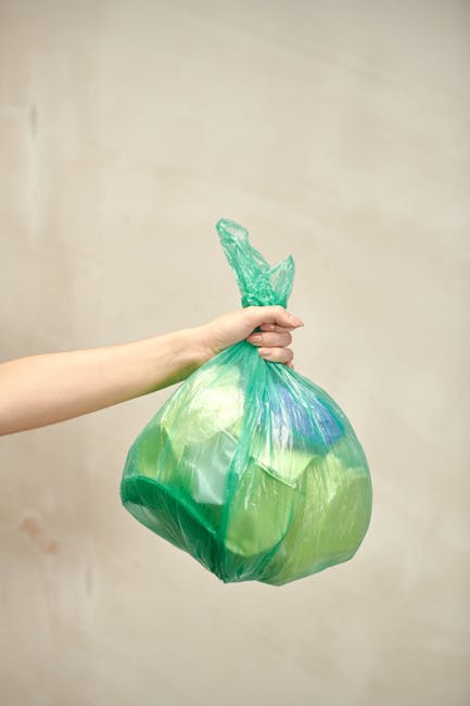 A person's hand is visible holding a green and blue plastic garbage bag, which appears to contain waste materials. The bag is tightly knotted at the top and held against a plain, light-colored wall ba
