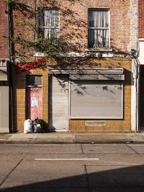 The image depicts the exterior of a commercial building with a weathered brick facade and two windows with partially drawn curtains on the upper floor. The ground level features a closed metal roller 